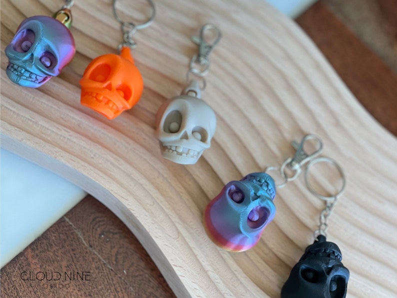 May include: Five colorful skull keychains with silver key rings. The skulls are made of plastic and have different colors and finishes, including orange, gray, black, and iridescent blue and purple.