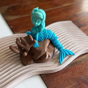 May include: A turquoise mermaid figurine with a blue tail sitting on a brown rock. The figurine is on a light brown, wavy surface.