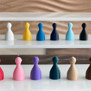 May include: A set of 12 game pieces in various colors, including red, pink, purple, teal, beige, brown, white, yellow, blue, dark blue, light blue, and black. The pieces are arranged in two rows on a white surface.