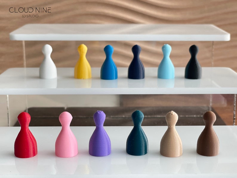 May include: A set of 12 game pieces in various colors, including red, pink, purple, teal, beige, brown, white, yellow, blue, dark blue, light blue, and black. The pieces are arranged in two rows on a white surface.