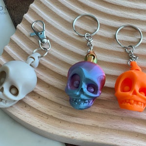 May include: Three skull keychains, one white, one iridescent blue and purple, and one orange. The keychains are on a wooden surface.