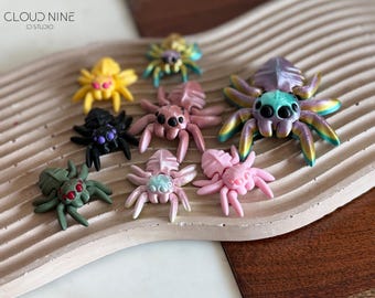 May include: A collection of nine colorful, hand-painted spider figurines. The spiders are made of polymer clay and have different colors, including pink, purple, green, yellow, and black.