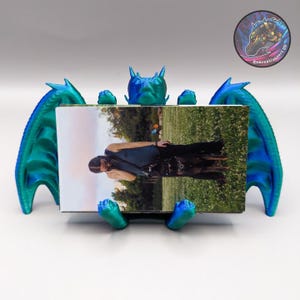 May include: A blue and green 3D printed dragon figurine holding a photo in its front paws. The dragon has wings and a spiky head. The photo shows a person standing in a grassy field.