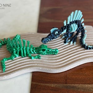 May include: Two 3D-printed dinosaur skeleton models. One is bright green, the other is black with teal accents. They are displayed on a light brown, wavy-patterned wooden base. The Cloud Nine 3D Studio logo is in the upper left corner.