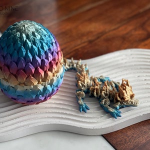 May include: A colorful dragon egg with iridescent scales and a matching dragon figurine with gold accents. The dragon is posed in a walking position with its head turned to the side. The egg and dragon are on a white textured surface.