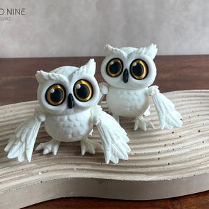 May include: Two white ceramic owl figurines with black eyes and gold accents. The owls are perched on a light brown wooden surface.