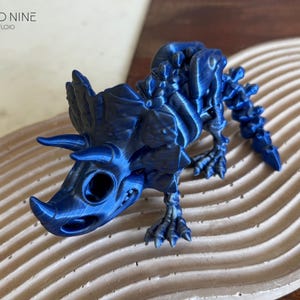 May include: A blue 3D-printed Triceratops skeleton model. The dinosaur figurine has a detailed design, including horns, a frill, and visible bone structure. The model is displayed on a textured, wavy surface. The Cloud Nine 3D Studio logo is in the upper left corner.