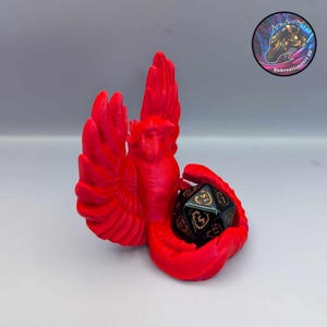 May include: A vibrant red, 3D-printed dice holder shaped like a stylized bird, cradling a black and gold twenty-sided die. The holder features outstretched wings and a detailed design. The die is approximately 1 inch in diameter. The logo "Rekreationsart 3D" is in the background.