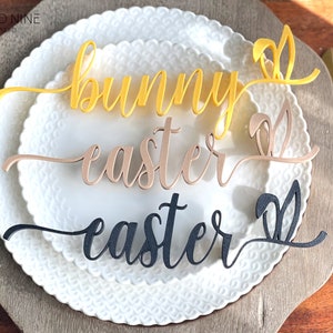 May include: Three wooden Easter place card holders with bunny ears. The words "bunny", "easter", and "easter" are written in cursive. The bunny ears are yellow, brown, and black.