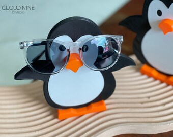 May include: A black and white penguin figurine wearing clear sunglasses with black lenses. The penguin is standing on an orange surfboard on a light brown surface. The text "CLOUD NINE 3D STUDIO" is visible in the top left corner of the image.