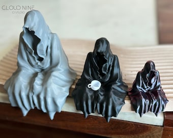 May include: Three 3D printed figurines of hooded figures, each in a different size and color. The largest is gray, the middle is black, and the smallest is purple. The middle figure is holding a small white cup.