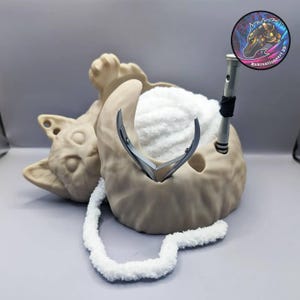 May include: A beige ceramic cat yarn bowl with a white yarn ball inside. The bowl has a small hole for a yarn needle and a slot for scissors. The cat is lying on its back with its paws in the air.