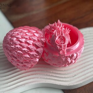 May include: A pink dragon figurine with rose details inside a pink egg-shaped container. The container is open and the dragon is visible. The container is made of a textured material that resembles scales.