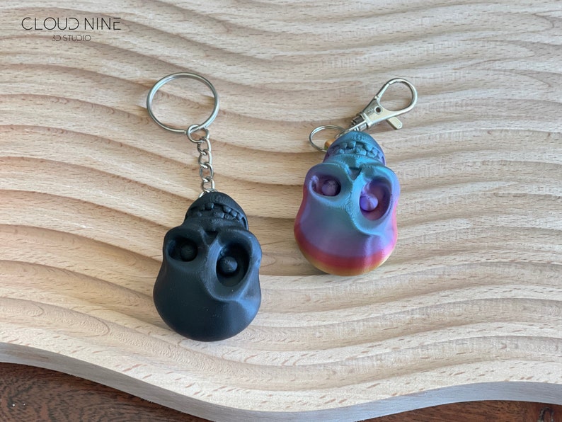 May include: Two skull-shaped keychains, one black and one with a rainbow gradient. Both keychains have silver metal rings.