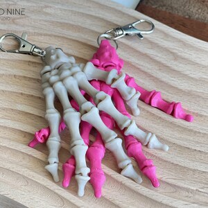 May include: Two skeleton hand keychains, one white and one pink, with silver key rings. The keychains are on a wooden surface.