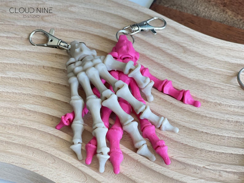 May include: Two skeleton hand keychains, one white and one pink, with silver key rings. The keychains are on a wooden surface.
