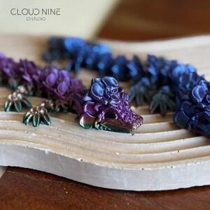 May include: A collection of articulated dragon figurines in shades of purple, blue, and green. The dragons are displayed on a light-colored, textured surface. The text "CLOUD NINE 3D STUDIO" is visible in the upper left corner.