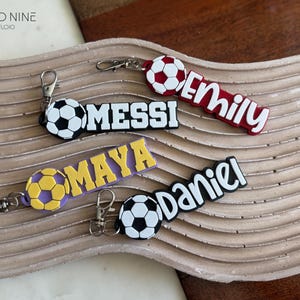 May include: Keychains with soccer ball designs and names: Messi, Emily, Maya, and Daniel. Each keychain has a soccer ball charm and a name tag in various colors, attached to a silver-tone clasp.