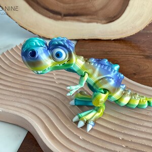 May include: A green, blue, and yellow 3D printed dinosaur toy with large eyes. The dinosaur is posed in a standing position with its head tilted to the side. The toy is sitting on a light brown surface.