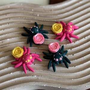 May include: Four small, handmade polymer clay spiders with pink and yellow flowers. The spiders have black bodies with white dots and pink legs. The flowers are yellow with pink centers.