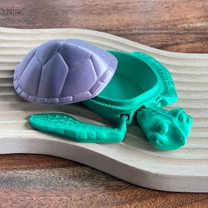 May include: A green turtle-shaped trinket box with a purple lid. The box is open and the lid is resting on a wooden surface.