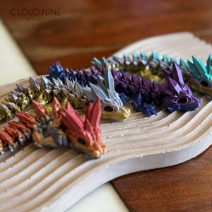May include: A collection of colorful, articulated dragon figurines. Each dragon features a detailed, crystalline design in shades of red, gold, purple, blue, and green. The dragons are displayed on a textured, light-colored surface.