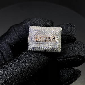 May include: A silver ring with a rectangular design featuring the word "SIMM" set with clear cubic zirconia stones.