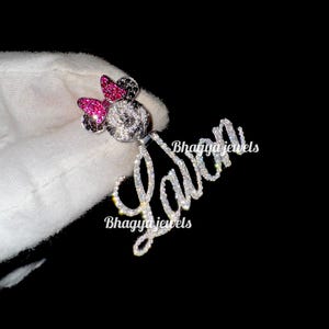 May include: A silver charm necklace with a sparkling diamond-encrusted name "Lavin" and a Mickey Mouse charm with pink bow and black ears. The charm is also covered in sparkling diamonds.