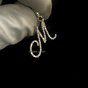 以下が含まれることがあります： ダイヤモンドをちりばめた「M」の文字のペンダント。ペンダントは黒い背景に保持されています。ペンダントは、金色の金属にセットされた、小さく輝くダイヤモンドでできています。ペンダントは繊細なジュエリーです。