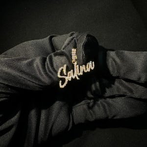 Op de afbeelding: Een goudkleurige hangerketting met de naam "Salina" in een cursief lettertype, ingelegd met kleine, sprankelende steentjes. De hanger hangt aan een delicate ketting. De ketting wordt tegen een donkere achtergrond gepresenteerd.