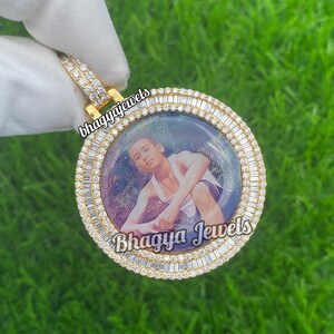 Pode incluir: Um pingente de ouro com um quadro de foto redondo cercado por uma dupla fileira de diamantes de corte baguette. A foto dentro do quadro mostra um jovem. O pingente &eacute; rotulado como "Bhagya Jewels".