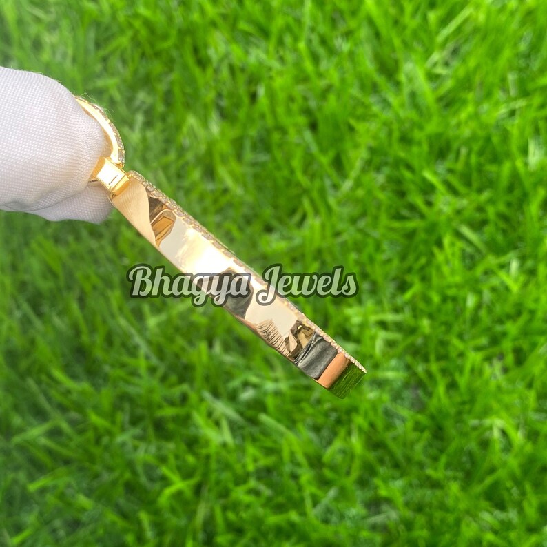 Pode incluir: Um bracelete dourado com uma borda redonda e chanfrada. O bracelete &eacute; adornado com pequenas pedras transparentes que criam um efeito brilhante. O bracelete &eacute; fotografado contra um fundo de grama verde. O texto "Bhagya Jewels" &eacute; vis&iacute;vel no bracelete.