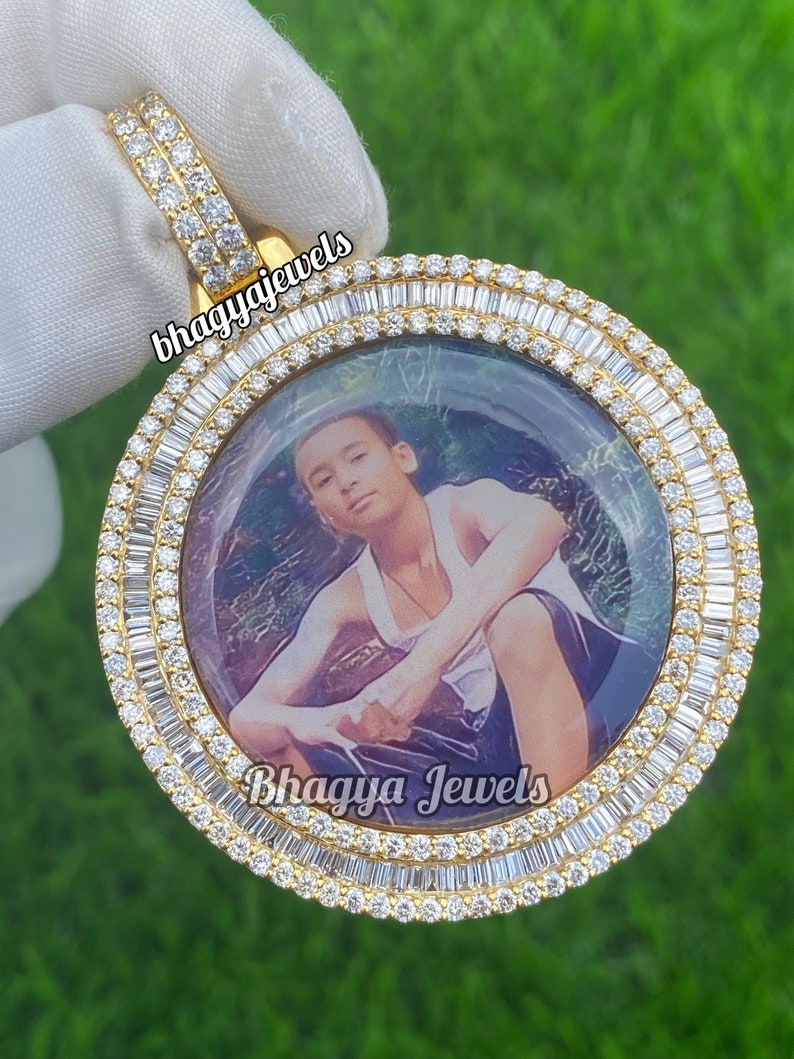 Pode incluir: Pingente de ouro com um quadro de foto redondo cercado por diamantes baguette. O quadro de foto tem uma imagem de um jovem. O pingente tem o texto "Bhagya Jewels" nele.