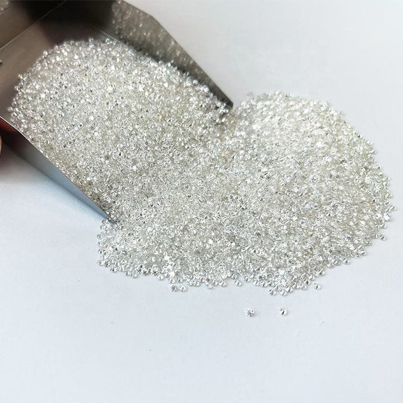 May include: A large scoop filled with loose, white diamonds. The diamonds are scattered on a white surface.