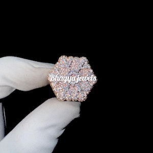 Könnte beinhalten: Ein großer, sechseckiger Ring mit einem blumenartigen Design. Der Ring ist mit klaren und rosafarbenen Steinen besetzt und hat eine Roségold-Oberfläche. Der Ring wird in einer behandschuhten Hand gehalten. Der Text "Bhagya Jewels" ist auf dem Ring sichtbar.