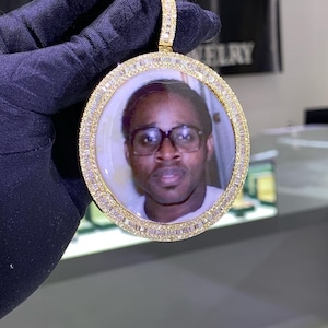 May include: A gold pendant featuring a round photo of a man wearing glasses. The pendant is encircled by a row of small diamonds.