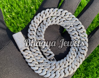 May include: A silver Cuban link chain necklace with a large, intricate design. The chain is covered in small, clear stones that give it a sparkling appearance. The chain is laid out on a black surface.