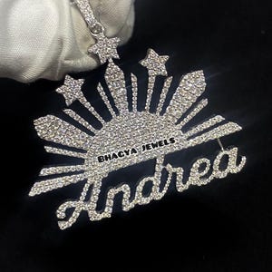 Könnte beinhalten: Silberner Anhänger mit dem Namen "Andrea" in Schreibschrift. Der Anhänger hat ein Sonnenstrahl-Design mit drei Sternen und dem Text "BHAGYA JEWELS". Der Schmuck ist mit kleinen, funkelnden Steinen besetzt.