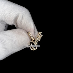 May include: A gold-colored pendant spelling out the name "Omar" in a cursive font, encrusted with small, sparkling gemstones. The pendant is held against a black background, with the brand name "Bhagya Jewels" visible.