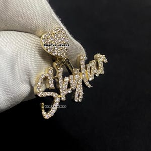 May include: A gold pendant spelling "Skylar" in a cursive font, embellished with small, sparkling stones. A heart-shaped design, also stone-covered, sits at the top. The pendant is held against a white cloth, with the brand name "BHAGYA JEWELS" visible.