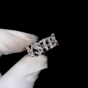 May include: A silver ring with the word "NSPIRE" spelled out in sparkling, clear gemstones. The ring is held against a black background, with the brand name "Bhagya jewels" visible. The ring is a piece of jewelry.