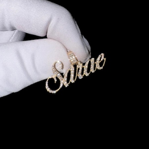 May include: A gold-colored pendant spelling out the name "Sarae" in a cursive font. The pendant is encrusted with small, clear gemstones. The pendant is held by a gloved hand against a black background. Jewelry.