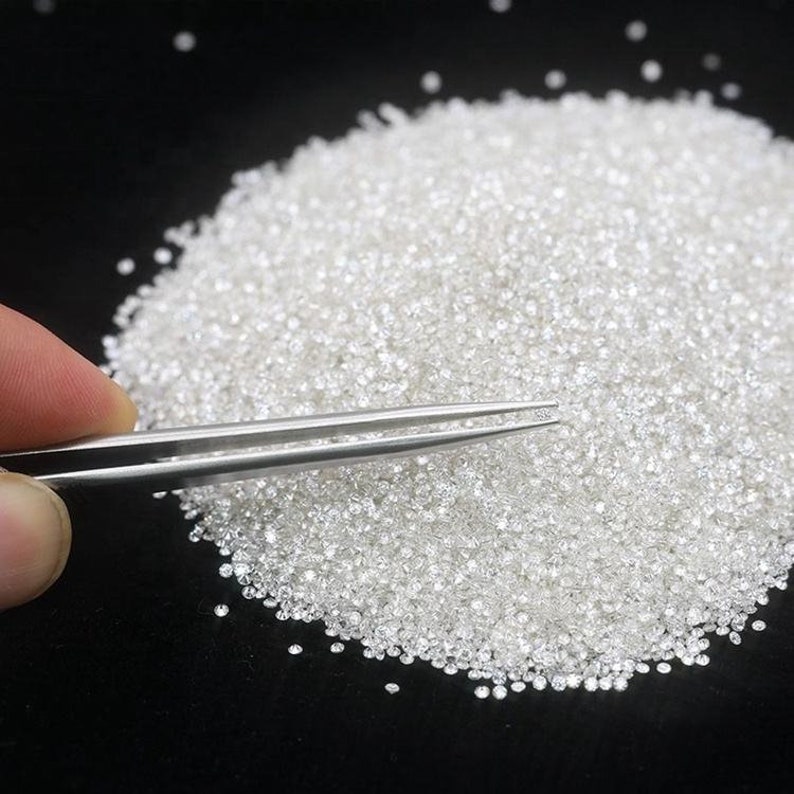 May include: A pile of small, white diamonds on a black surface. A pair of tweezers holds a single diamond.