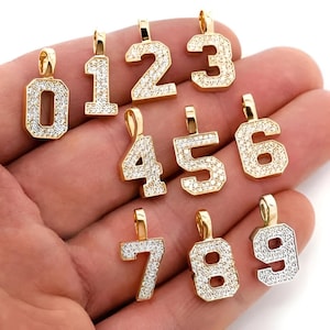 May include: Ten gold-tone pendants, each featuring a different number from 0 to 9, are set with clear cubic zirconia stones. The pendants are displayed on a hand.
