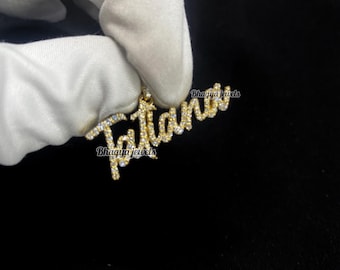Puede incluir: Un colgante dorado con el nombre "Tatiana" escrito en cursiva, engastado con pequeñas piedras preciosas transparentes. El colgante se sostiene sobre un fondo oscuro. También es visible el nombre de la marca "Bhagya jewels".