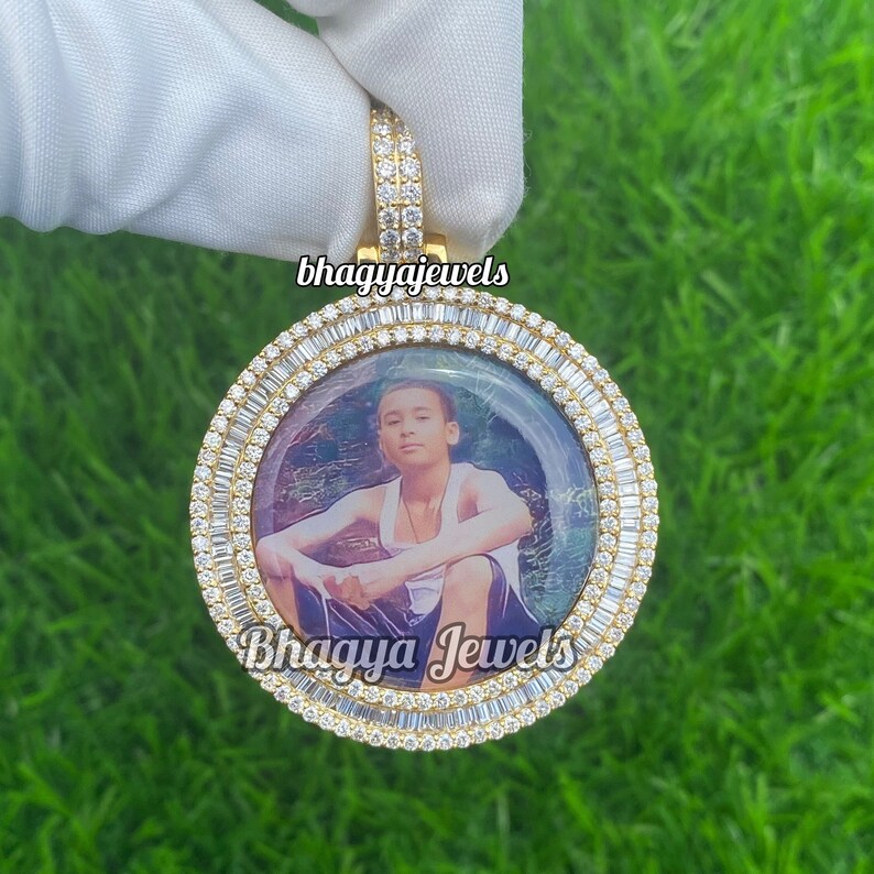 Pode incluir: Um pingente de ouro com um quadro de foto redondo cercado por diamantes baguette. A foto dentro do quadro mostra um jovem vestindo uma camiseta branca sem mangas e sentado em um banco. O pingente tem o texto "Bhagya Jewels" na parte inferior.