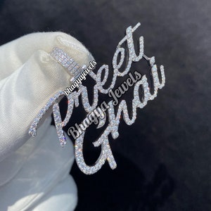 Può includere: Un ciondolo d'argento con il nome "Preet Ghai" in corsivo, incastonato con piccole pietre scintillanti. Il ciondolo è tenuto da una mano guantata, con il testo "Bhagya Jewels" visibile.