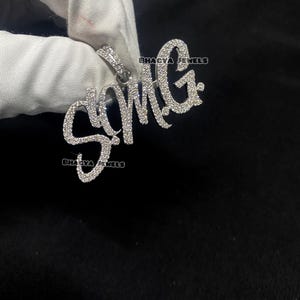 Może przedstawiać: Zawieszka wysadzana diamentami z napisem "SMG" w kursywie. Zawieszka jest trzymana na czarnym tle, z widocznymi słowami "BHAGYA JEWELS". Zawieszka wykonana jest z małych, błyszczących kamieni.