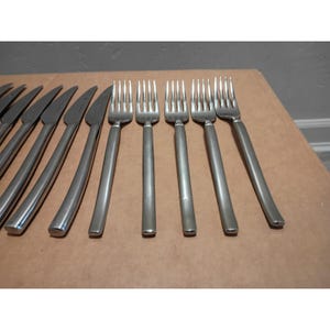 May include: A set of stainless steel flatware consisting of five forks and six knives arranged in two rows. The forks feature four tines and long, smooth handles, while the knives have curved blades and matching metallic handles with a polished finish.
