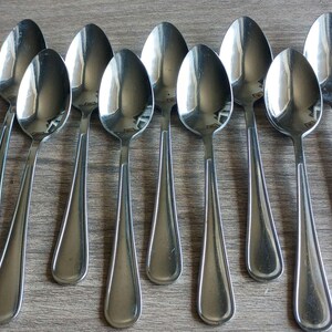 May include: A set of eight polished stainless steel spoons arranged in two rows. Each spoon features a smooth, rounded bowl and a simple, tapered handle with a subtle raised line detail running down the center of the stem, reflecting light.